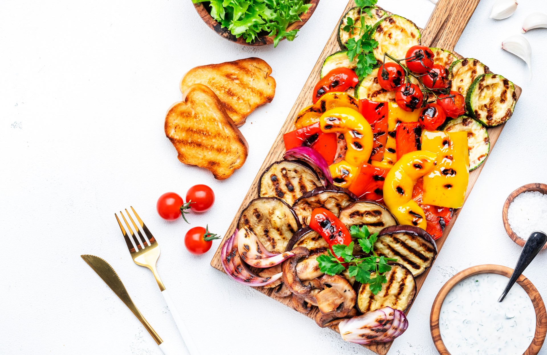 Vegan lunch with grilled vegetables: red and yellow paprika, zucchini, eggplant, mushrooms, tomatoes and red onion served on rustic wooden cutting board, white table background, top view